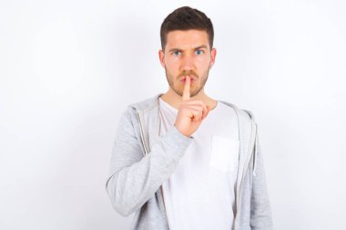 Beyaz arka plan üzerine gündelik kıyafetler giyen genç beyaz bir adam sessiz ol diyor. Sırrımı kimseye söyleme ya da yüksek sesle konuşma, lütfen.!