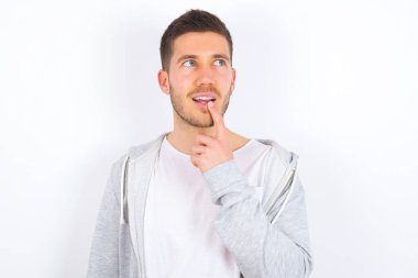 Beyaz arka plan üzerine günlük giysiler giymiş genç beyaz bir adam düşünceli bir ifadeyle kameraya bakıyor, elini yüzüne yaklaştırıyor, parmağını ısırıyor hoş bir şey düşünüyor..