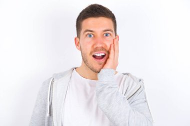Şok olmuş, şaşkın, beyaz arka plan üzerine günlük giysiler giyen beyaz bir adam ağzı açık, ağzı açık, eli yüzün yanında şaşırmış görünüyor. Olumlu duygu yüz ifadesi vücut dili.