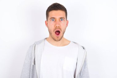 Etkileyici yüz ifadeleri. Şok olmuş, beyaz arka planda günlük kıyafetler giyen, ağzı açık gezen genç beyaz adam gördüklerinden sersemlemiş hissediyor..