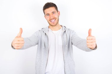 Beyaz arka plan üzerine günlük kıyafetler giyen genç beyaz bir adam olumlu bir jest yapıyor. Başparmağıyla gülümsüyor ve başarı için seviniyor. Kameraya bak, kazanan jest..