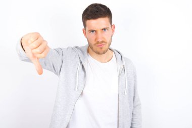 Beyaz arka plan üzerine günlük kıyafetler giyen hoşnutsuz genç beyaz bir adam onaylanmama işareti gösteriyor, baş parmağını aşağıda tutuyor, hoşnutsuzluğunu dile getiriyor, hoşnutsuzluğunu ifade ediyor. Olumsuz duygular.