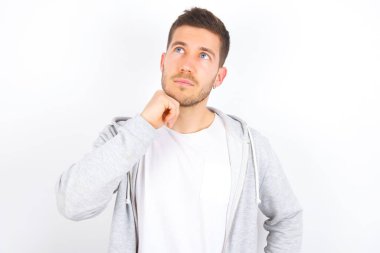 Yüz ifadeleri ve duygular. Düşünceli, beyaz arka plan üzerine günlük giysiler giymiş genç beyaz bir adam başının altında el ele tutuşmuş, şüpheli bir bakışı var..