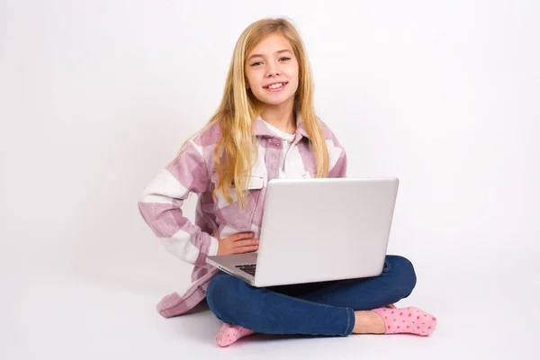 スタジオショットの陽気な美しいですCaucasianティーン女の子座っていますノートパソコンで蓮の位置に白い背景に手を維持します 笑顔広く — ストック写真