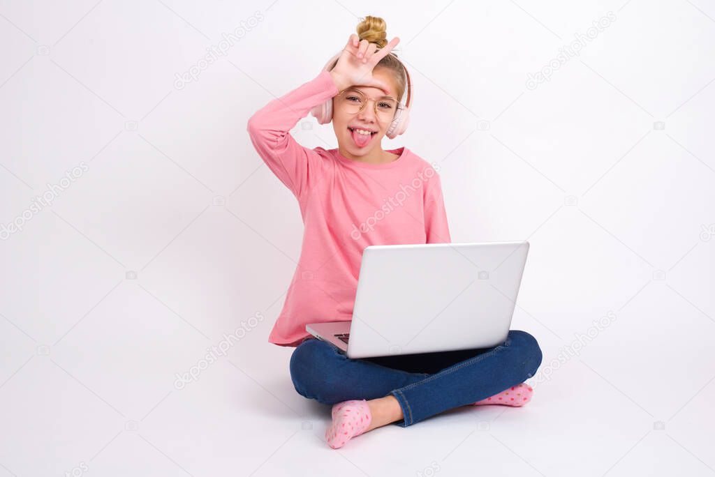 Funny Beautiful caucasian teen girl sitting with laptop in lotus position on white background makes loser gesture mocking at someone sticks out tongue making grimace face.