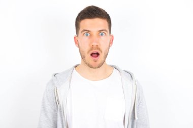 Duygusal çekiciliği olan beyaz arka plan üzerine günlük kıyafetler giyen beyaz tenli genç bir adam ağzı açık bir şekilde büyük bir şaşkınlık ve korku içinde kameraya bakıyor. Beklenmedik haberler ve insani tepkiler.