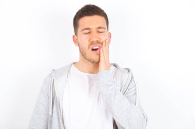 Diş ağrısı konsepti. Beyaz arka plan üzerine günlük kıyafetler giyen genç beyaz adam acı hissediyor, yanağını eliyle tutuyor, kötü diş ağrısından muzdarip, acı dolu bir ifadeyle kameraya bakıyor.