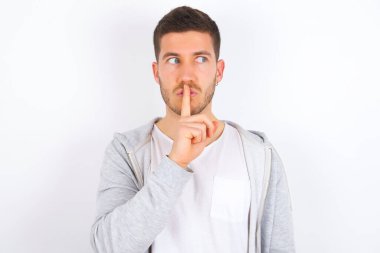 Beyaz arka plan üzerine günlük kıyafetler giyen genç beyaz bir adam sessiz bir jest yapar, işaret parmağını dudaklara tutar ve sessiz olur. Sırrımızı paylaşmamamızı istiyor..