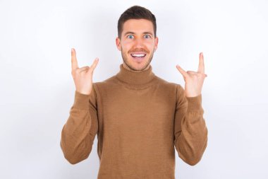 Beyaz arka plan üzerine balıkçı yaka giyen genç beyaz adam rock 'n roll tabelasını kendinden emin ve neşeli gösteriyor. Partide havalı müzikten hoşlanıyor. Vücut dili kavramı.