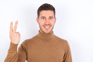 Beyaz arka plan üzerine balıkçı yaka giyen genç beyaz bir adam gülümsüyor ve arkadaş canlısı görünüyor, iki ya da iki numara önde, geriye doğru sayıyor