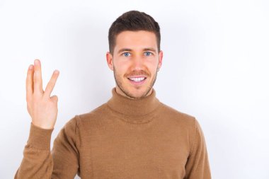 Beyaz arka plan üzerine balıkçı yaka giyen genç beyaz bir adam gülümsüyor ve arkadaş canlısı görünüyor, üç ya da üç numarayı gösteriyor, geriye doğru sayıyor.