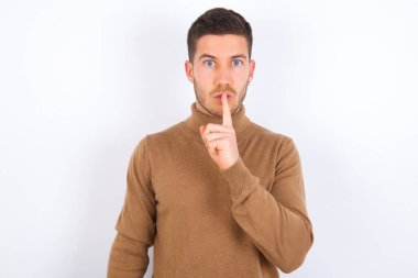 Şaşırmış genç beyaz adam sessiz bir jest yapıyor, parmaklarını dudaklarının üstünde tutuyor ve kameraya gizemli bakıyor.