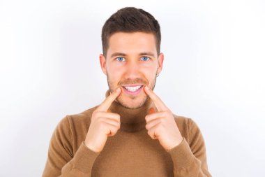 Beyaz arka plan üzerine balıkçı yaka giyen mutlu, genç beyaz tenli, dişlek gülüşlü, işaret parmakları ağızda, parmakları işaret eden ve neşeli gülümsemeye zorlayan bir adam.