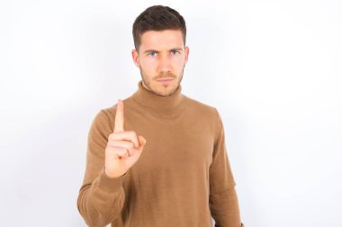 Beyaz arka plan üzerine balıkçı yaka giyen genç beyaz adam hayal kırıklığına uğramış ve ön tarafı işaret ediyor.