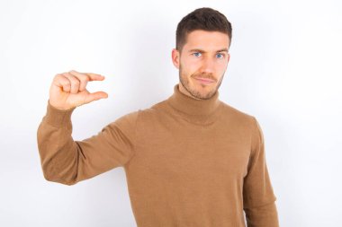 Beyaz arka plan üzerine balıkçı yaka giyen hoşnutsuz genç beyaz adam küçük el işaretleri çok büyük olmayan bir şeyi gösteriyor. Vücut dili kavramı.