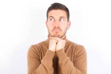 Beyaz arka plan üzerine balıkçı yaka giyen meraklı genç beyaz adam ellerini çenesinin altında tutuyor dudaklarını ısırıyor ve ilgiyle bakıyor..