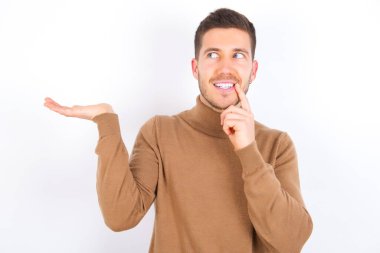 Beyaz arka plan reklamı üzerine balıkçı yaka giyen olumlu genç beyaz adam parmak ile dişlere dokunuyor..