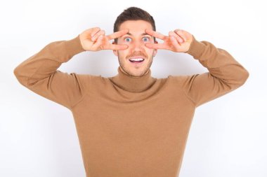 Beyaz arka planda balıkçı yaka kazak giyen neşeli, pozitif genç beyaz bir adam, gözlerin yanında V işareti gösteriyor.