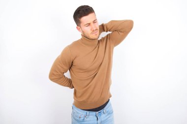 Beyaz arka plan üzerine balıkçı yaka giyen genç beyaz bir adam stüdyoda sırt ağrısı çekti.