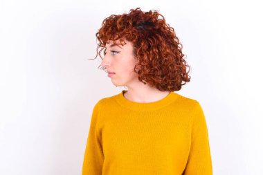 Beyaz arka planda sarı süveter giyen, gülümseyen genç, kızıl saçlı kızın yan görüntüsü.  