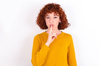 Beyaz arka planda sarı süveter giyen genç kızıl kız sessiz ol diyor. Sırrımı kimseye söyleme ya da yüksek sesle konuşma, lütfen.!