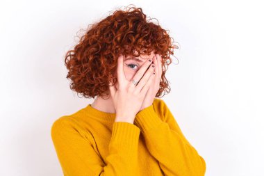 Beyaz arka planda sarı süveter giyen genç kızıl kız elleriyle yüzünü kapatıyor ve bir gözü parmaklarının arasında dışarı bakıyor. Bir şeyden ya da birinden korktun mu?.