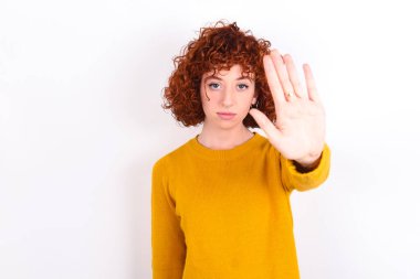 Beyaz arka planda sarı süveter giyen genç kızıl kız avuç içiyle el hareketi yapıyor. Yüzünde olumsuz ve ciddi bir jest olan uyarı ifadesi.