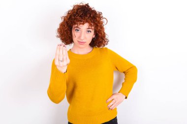 What the hell are you talking about. Shot of frustrated young redhead girl wearing yellow sweater over white background   gesturing with raised hand doing Italian gesture, frowning, being displeased and confused with dumb question.