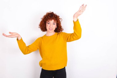 Beyaz arka planda sarı süveter giyen neşeli, genç kızıl kız hoş geldin jesti yapıyor ve kollarını başının üstüne kaldırıyor..