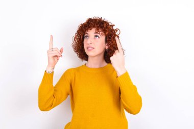 Başarılı, dost canlısı, kızıl saçlı, sarı süveter giyen, beyaz arka planda heyecanla bağıran, her iki işaret parmağıyla işaret eden,.