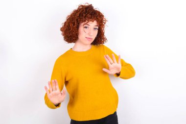 Beyaz arka planda sarı süveter giyen, korkmuş genç, kızıl saçlı bir kız, dehşete kapılmış bir ifade takınıyor ve iki elinle hareket etmeyi kes, orada kal. Panik konsepti.