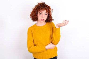Studio shot of frustrated young redhead girl wearing yellow sweater over white background   gesturing with raised palm, frowning, being displeased and confused with dumb question.