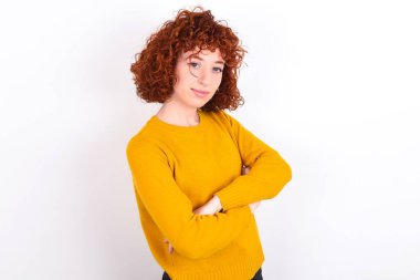 Beyaz arka planda sarı süveter giyen, kollarını kavuşturmuş neşeli kızıl saçlı bir kız resmi. Kameraya bakıp gülümsüyor. Güven kavramı.