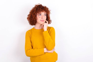 Yüz ifadeleri ve duygular. Beyaz arka plan üzerine sarı süveter giyen düşünceli genç bir kız başının altından elini tutuyor, şüpheli bir bakışı var..