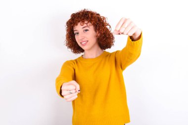 Beyaz arka planda sarı süveter giyen, el ele tutuşurken geniş bir gülümsemeyle gülümseyen çekici kızıl saçlı kızın portresi. Güven ve rahatlama kavramı.