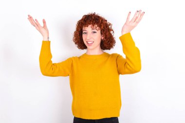 Neşeli, şanslı, sarı süveter giyen, beyaz arka planda tezahürat yapan, başarıyı kutlayan, yumruklarını sıkarak evet diye bağıran genç, kızıl saçlı kız.