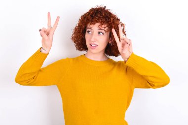 Beyaz arka planda sarı süveter giyen neşeli, genç bir kızın izole edilmiş fotoğrafı iki elinle barış ya da zafer işareti yapar..