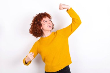 Beyaz arka planda sarı süveter giyen çekici genç bir kızıl kız zaferi kutluyor yumruklarıyla havaya yumruk atıyor ve ışıltılı bir gülümsemesi var.