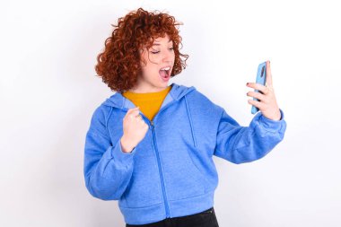 Beyaz arka plan üzerine mavi ceket giyen mutlu, genç kızıl kız e-posta yoluyla iyi haberler alıyor ve ayakta durup cep telefonuna bakarken başarıyı kutluyor..