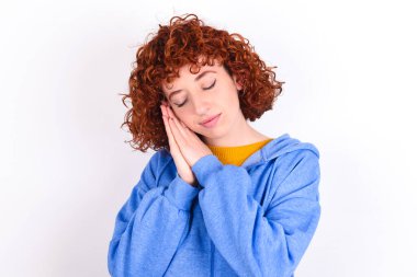 Beyaz arka planda mavi ceket giyen genç kızıl kız avuçlarına yaslanıyor gözlerini kapıyor ve hoş bir gülümseme görüyor.