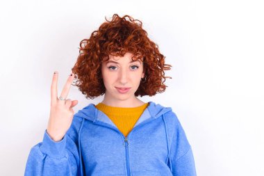 Beyaz arka planda mavi ceketli genç bir kız gülümsüyor ve arkadaş canlısı görünüyor, iki ya da iki numarayı gösteriyor, geriye doğru sayıyor.