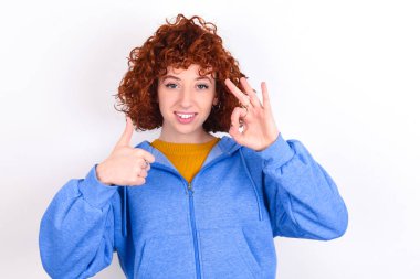 Beyaz arka planda mavi ceketli genç bir kız gülümsüyor ve mutlu görünüyor, gamsız ve pozitif, bir eliyle zafer ya da barış işareti yapıyor.