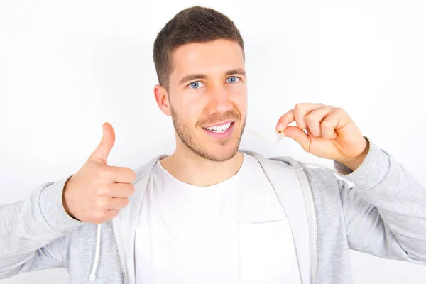 Beyaz arka plan üzerine günlük kıyafetler giyen genç beyaz bir adam görünmez bir diş teli tutuyor ve baş parmağını kaldırıyor, bu yeni tedaviyi öneriyor. Diş sağlığı konsepti.