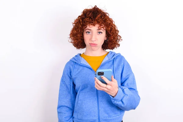 Расстроенная Рыжая Девушка Синей Куртке Белом Фоне Использует Мобильное Программное — стоковое фото
