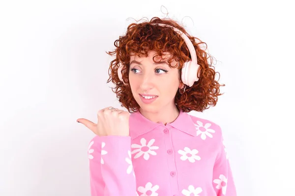 年轻的红头发女孩穿着粉色花T恤 背景为白色 通过无线耳机听音频 指尖远离广告复制空间 — 图库照片