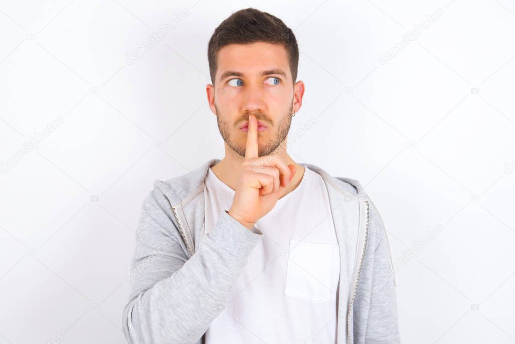 young caucasian man wearing casual clothes over white background makes silence gesture, keeps index finger to lips makes hush sign. Asks not to share secret.