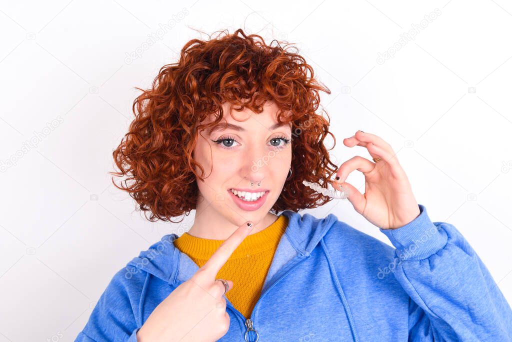 young redhead girl wearing blue jacket over white background holding an invisible aligner and pointing to her perfect straight teeth. Dental healthcare and confidence concept.