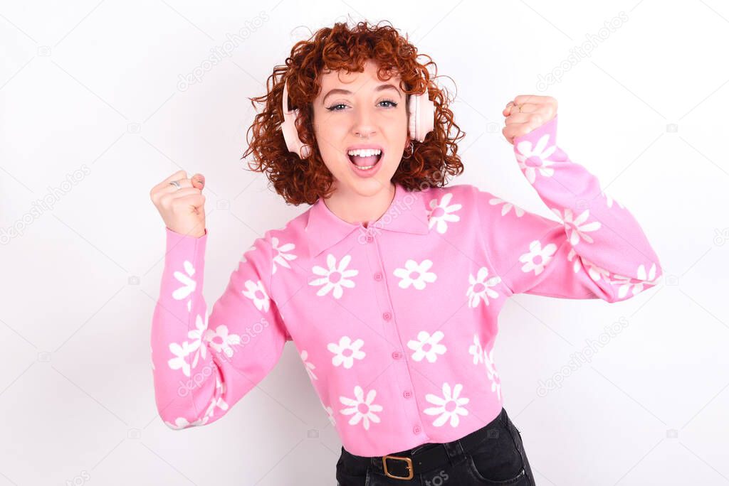 Emotional young redhead girl wearing pink floral t-shirt over white background exclaims loudly feels like winner raises clenched fists keeps mouth opened wears stereo headphones on ears makes yes gesture, listens favourite music