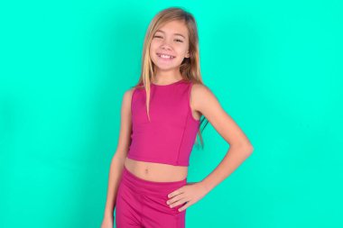 Kafkasyalı küçük neşeli kızın stüdyo fotoğrafı elini kalçasında tutuyor, geniş bir gülümsemeyle..