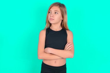 Yeşil arka planda siyah spor giysiler giyen düşünceli, düşünceli, sarışın küçük bir kız. Kollarını kavuşturmuş, düşünceli bir ifadeyle ne yapacağını düşünüyor.